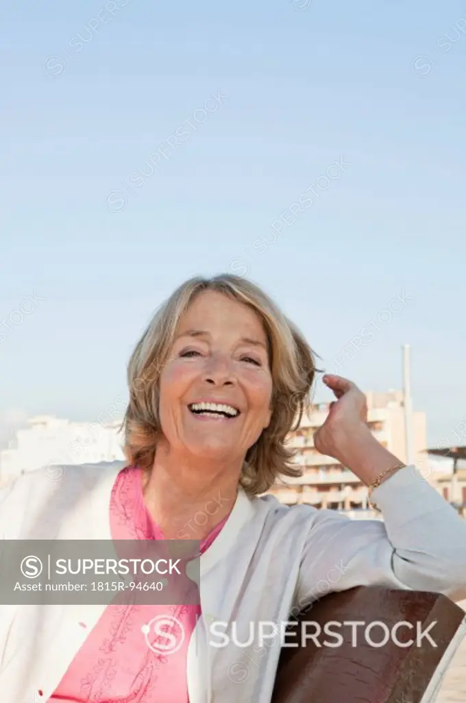 Spain, Mallorca, Senior woman sitting on bench at sea shore, smiling, portrait