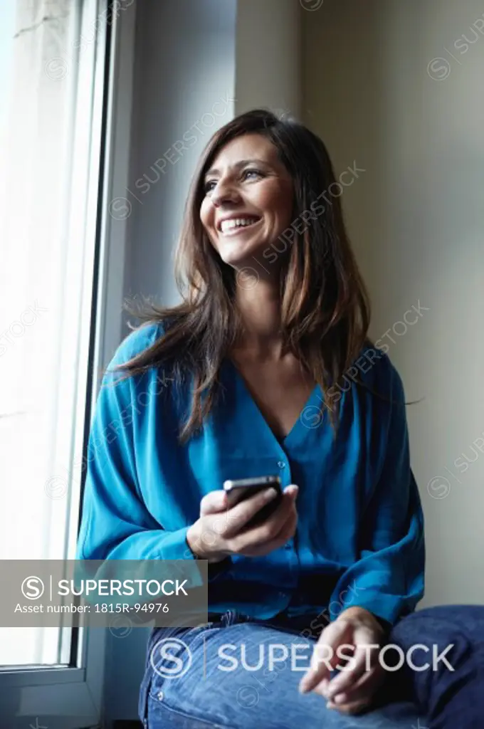 Germany, Cologne, Mid adult woman at window with cell phone, smiling