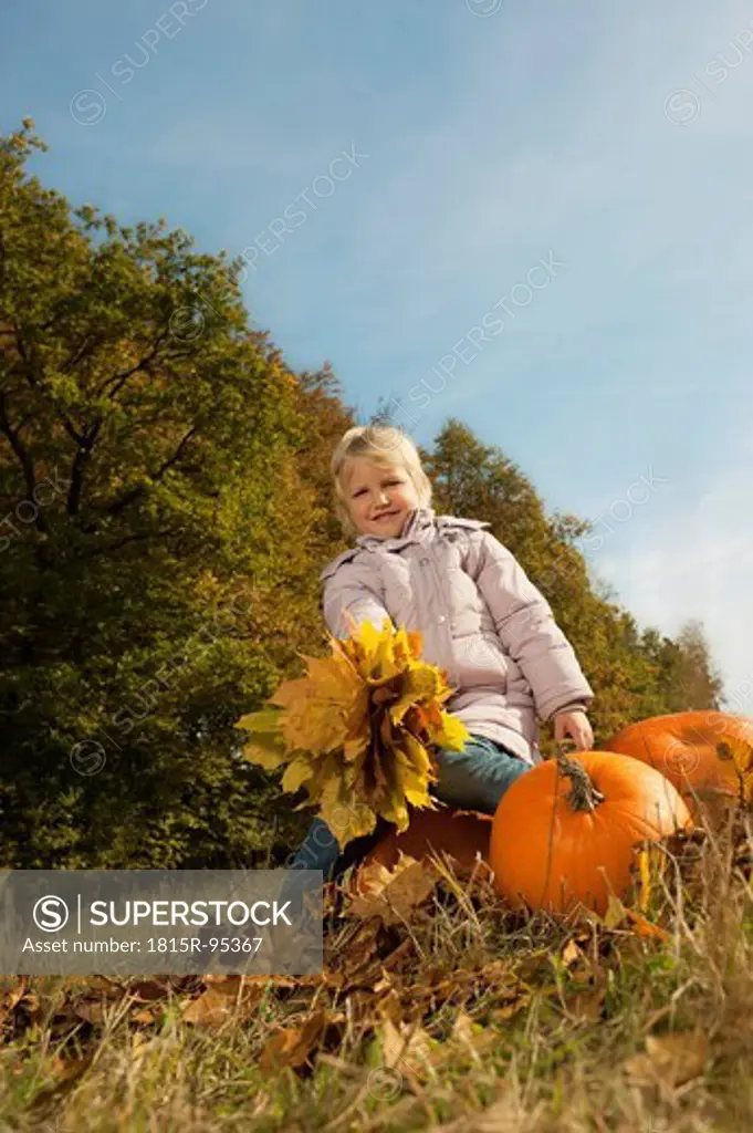 Germany, Bavaria, Girl sitting on pumpkin with leaves, smiling, portrait