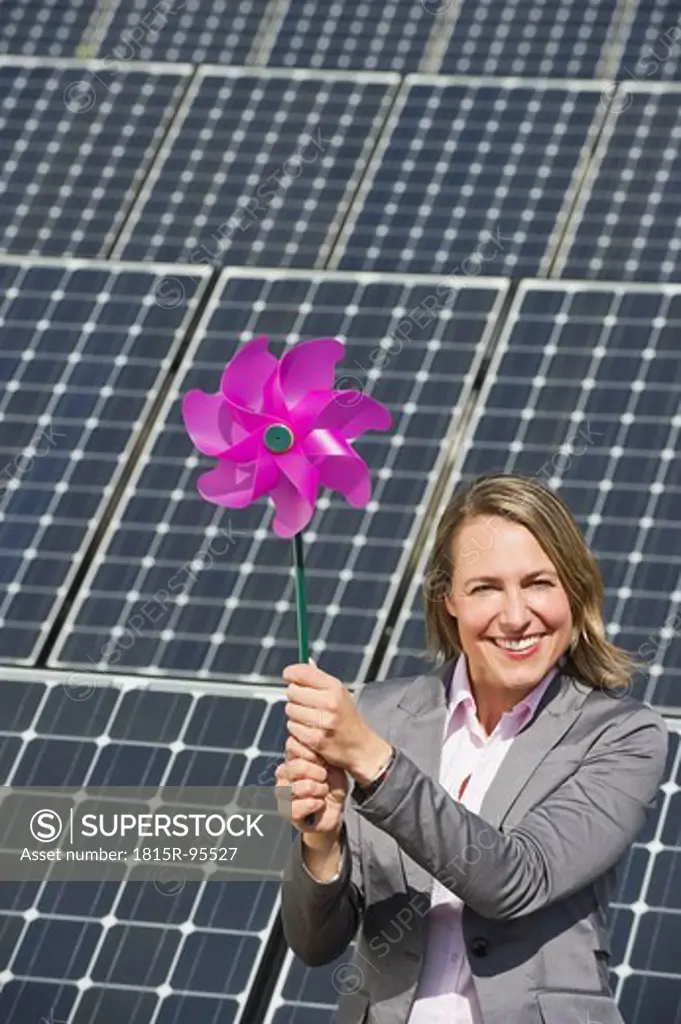 Germany, Munich, Woman holding paper windmill against solar panels, smiling, portrait