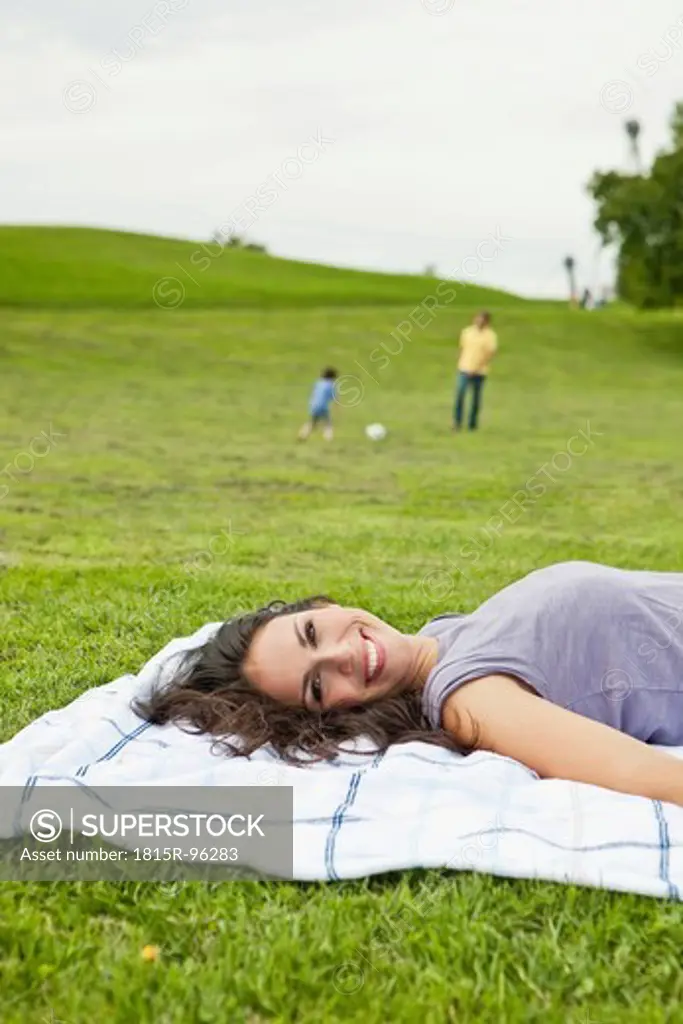 Germany, Bavaria, Woman lying on blanket, father and son playing soccer in park, smiling