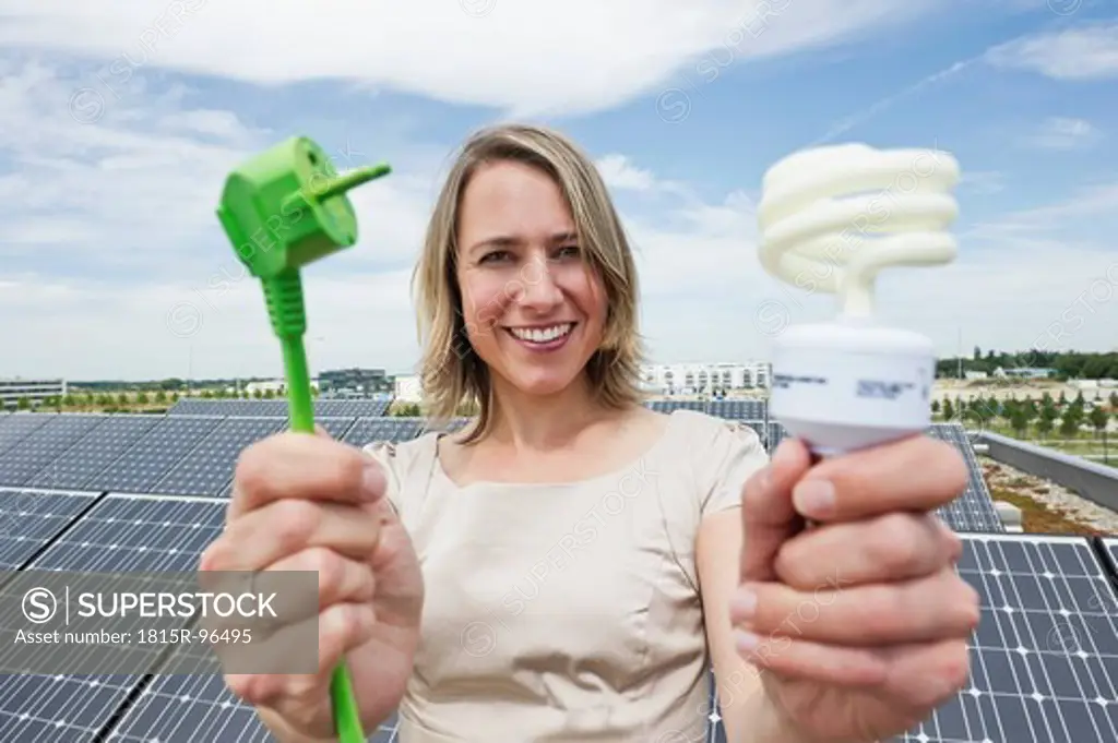 Germany, Munich, Woman holding electric cord and lightbulb in solar energy, smiling, portrait