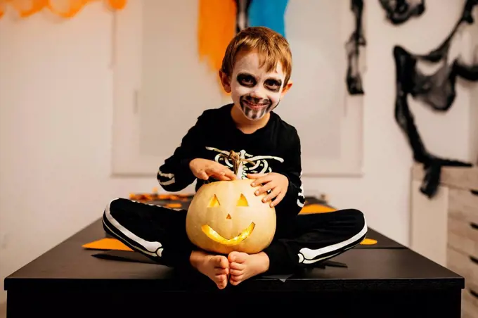 Portrait of smiling little boy with painted face and fancy dress sitting on table with Jack O'Lantern