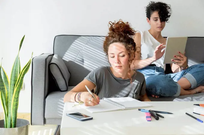 Young woman taking notes at home with friend sitting on couch using tablet