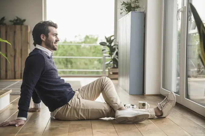 Young man sitting on floor, looking out of window, relaxing