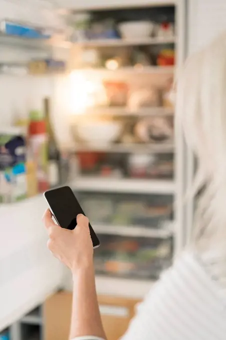 Hand of woman with smartphone checking fridge in kitchen at smart home