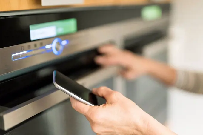Hand of woman with smartphone checking oven in kitchen at her smart home