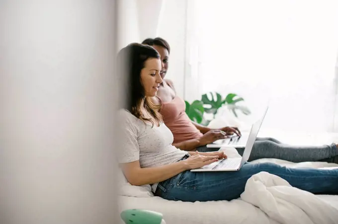 Two women lying in bed at home using laptops