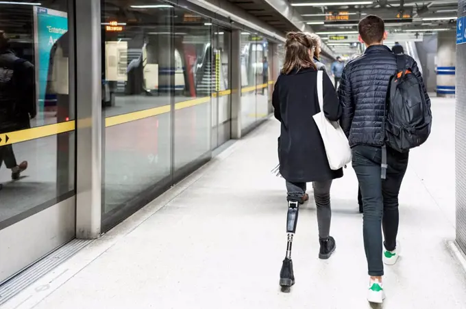 Rear view of young woman with leg prosthesis and man walking at subway station platfom