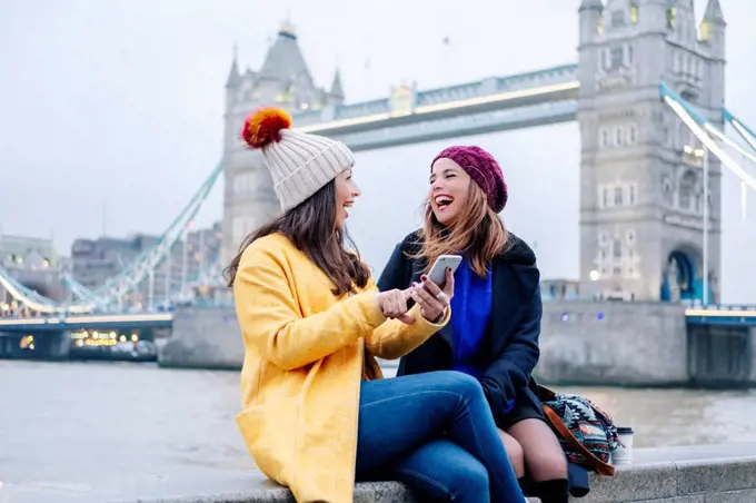 Two young tourists sitting on wall, using smartphone, with London Bridge in background
