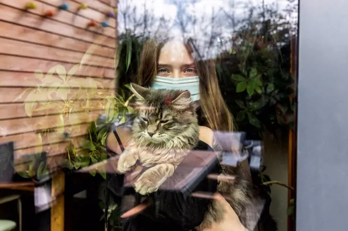 Portrait of girl with surgical mask and cat behind window pane