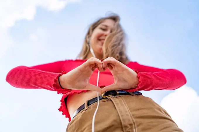 Worm's eye view of young woman listening to music with earphones shaping a heart with her hands