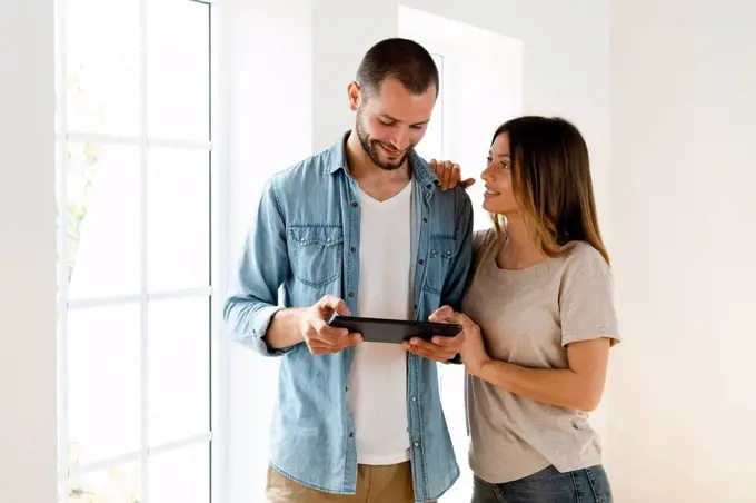 Smiling couple at home standing in front of window looking at tablet together