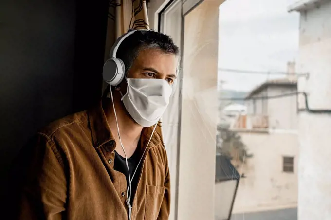Man wearing protective mask and white headphones looking out of the window
