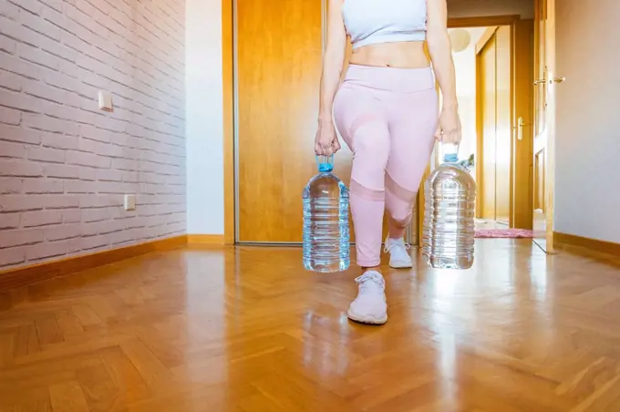 Low section of woman practicing lunges with water bottles during weight training at home