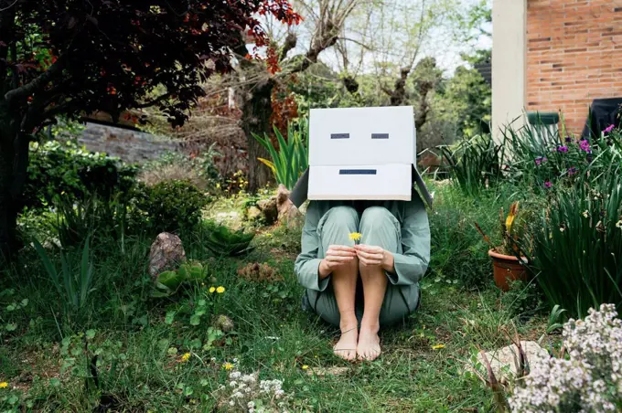 Young woman with cardboard box on her head sitting barefoot in garden