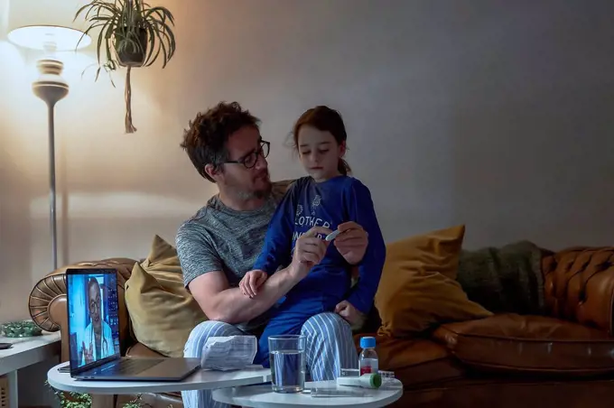 Man holding thermometer while sitting with sick daughter during video call at home