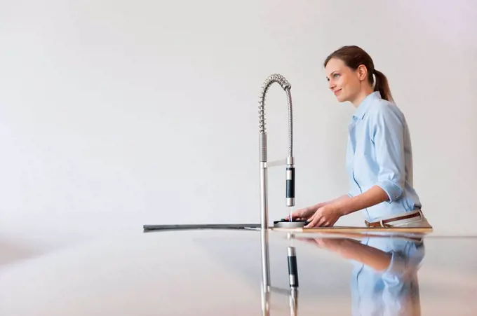 Smiling woman filling water in container through modern kitchen faucet against wall