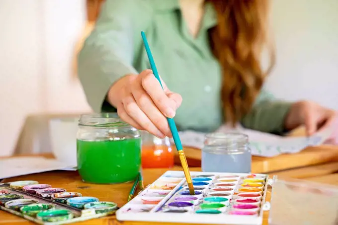 Young woman dipping paintbrush in watercolor on table at home