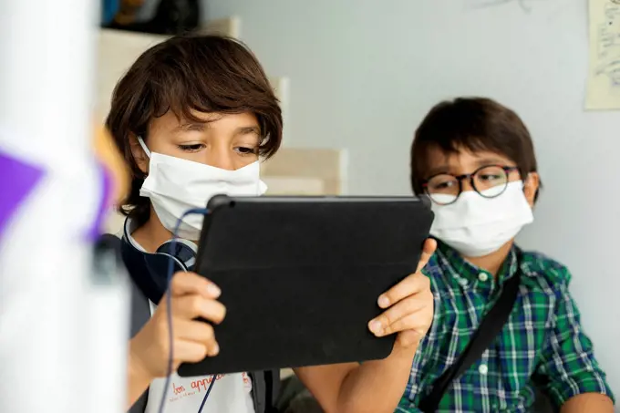 Boy wearing face mask using digital tablet while sitting in distance with friend at school