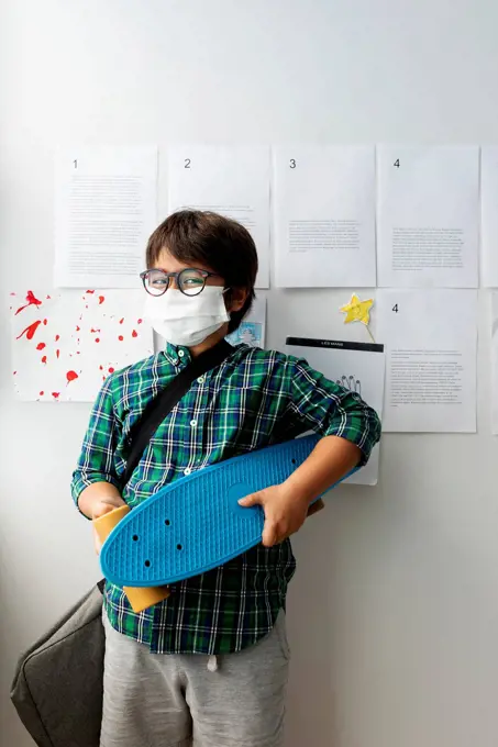 Boy wearing mask holding skateboard while standing against wall in school