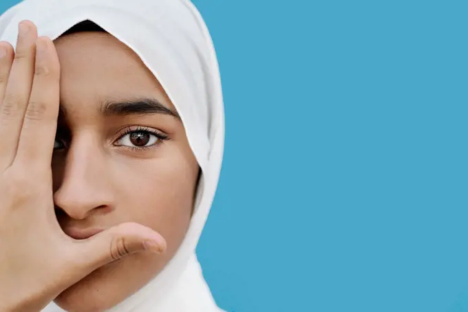 Muslim girl covering eye with hand against blue background