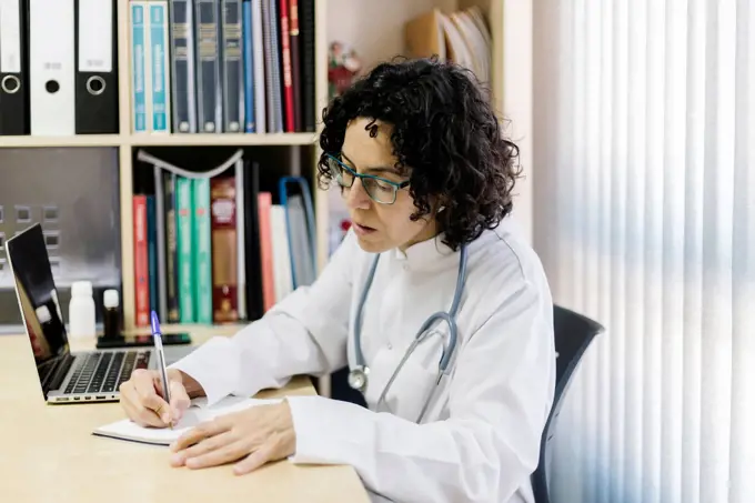 Female doctor writing by laptop while sitting at desk in office