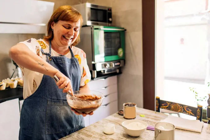 Smiling female baker mixing cake batter in bowl on table at workshop