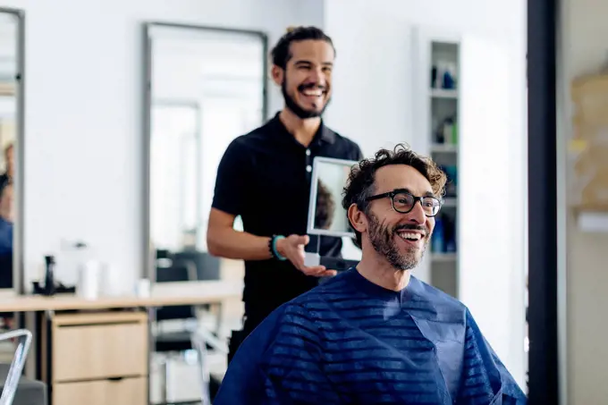Happy hairdresser showing haircut to client in mirror at salon
