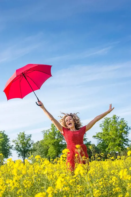 Carefree woman with arms raised holding umbrella while standing amidst oilseed rapes