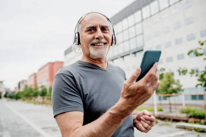 Smiling senior man wearing headphones using smart phone while standing in city
