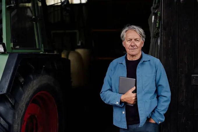 Portrait of a confident senior man holding tablet on a farm with tractor in barn