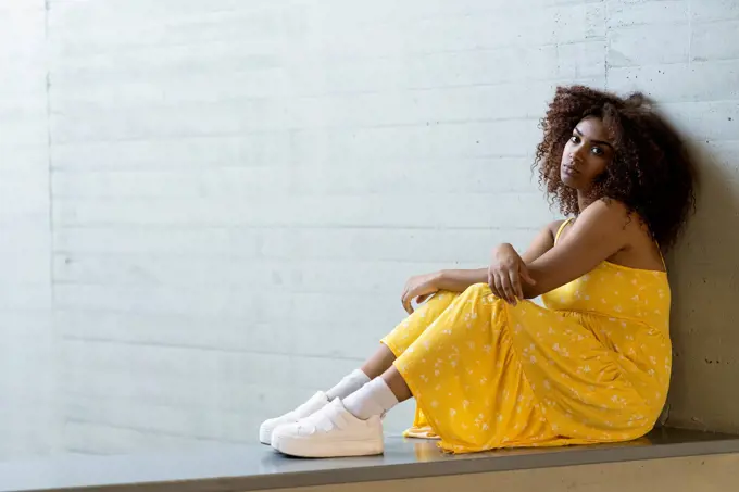 Sad woman with afro hair wearing yellow dress sitting on retaining wall