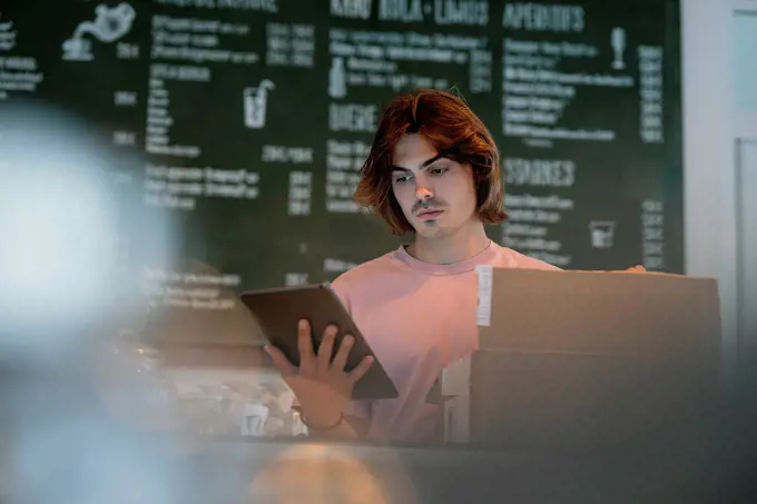 Close-up of serious male owner with box on counter using digital tablet in cafe