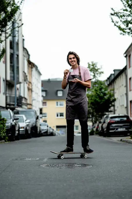 Smiling male owner holding coffee cup while skateboarding on road in city