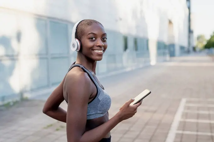 Smiling young woman wearing headphones using smart phone while standing in city