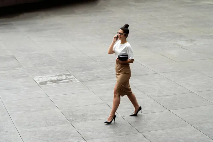 Young businesswoman talking through mobile phone while walking on footpath in city