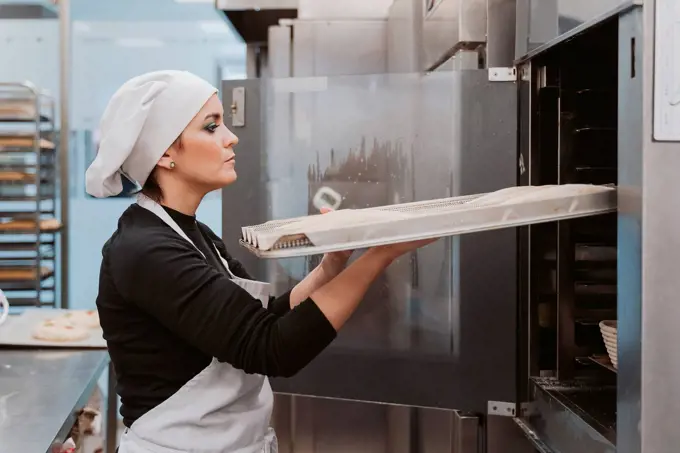 Female baker inserting baking tray in oven at bakery