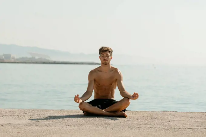 Shirtless young man meditating while sitting against sea at harbor