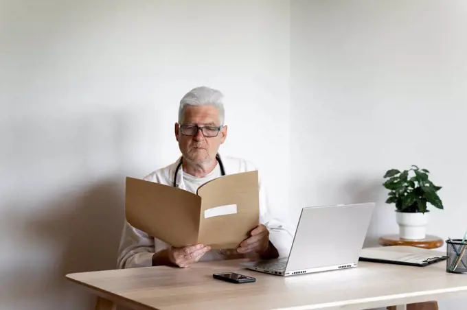 Senior doctor examining medical record while sitting at desk in clinic