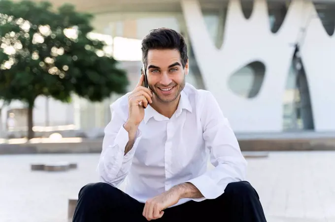 Smiling male entrepreneur talking over smart phone sitting against built structure in city