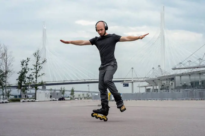Young man with arms outstretched listening music while inline skating on road against sky
