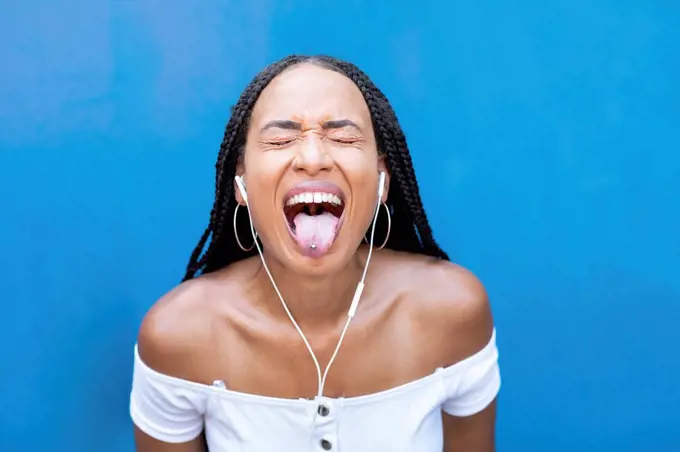 Woman listening music while sticking tongue out against blue wall