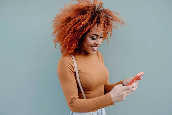 Young woman with curly hair using mobile phone while standing by wall