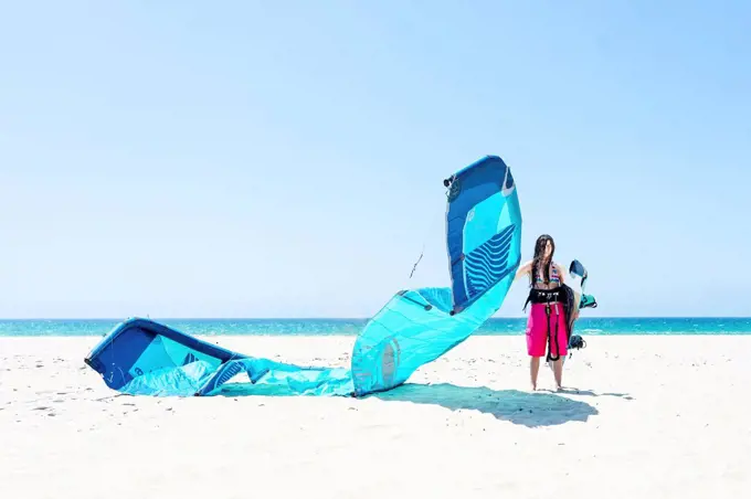 Young woman with kite and kiteboard standing at beach against clear sky during sunny day