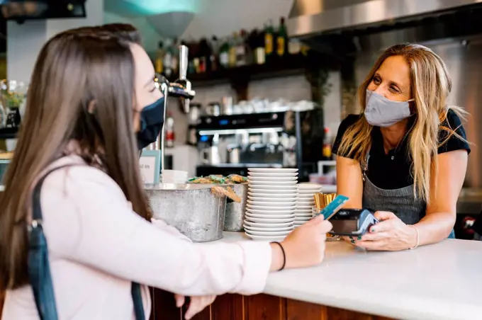 Businesswoman in face mask paying through credit card at counter in cafe during COVID-19 crisis