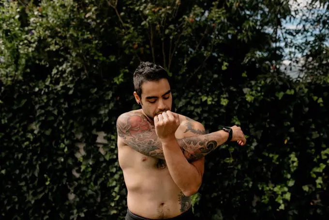 Shirtless handsome man with tattoo stretching arm while doing warm up exercise against plants at back yard