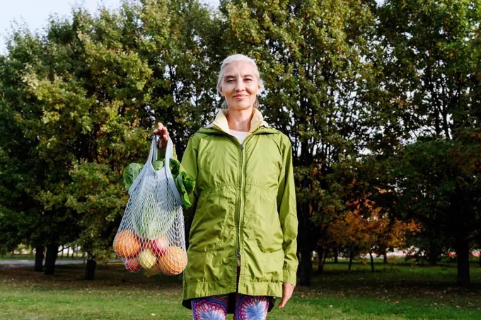 Mature woman holding mesh bag filled with fruits and vegetables at public park