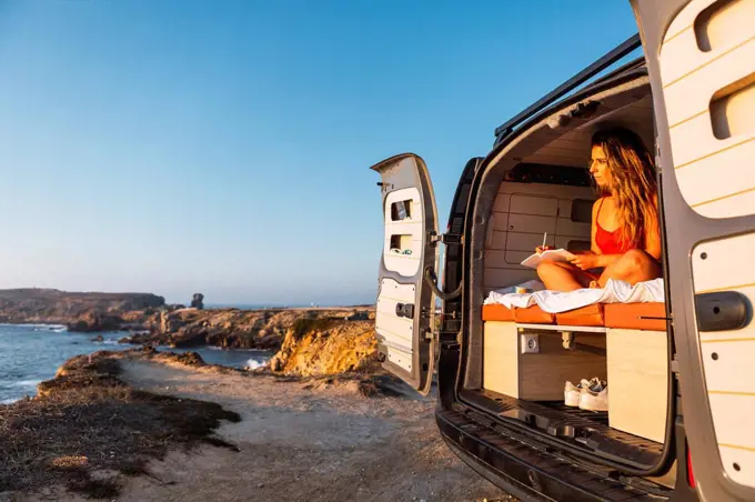 Young woman writing in book while sitting in camper van against at beach