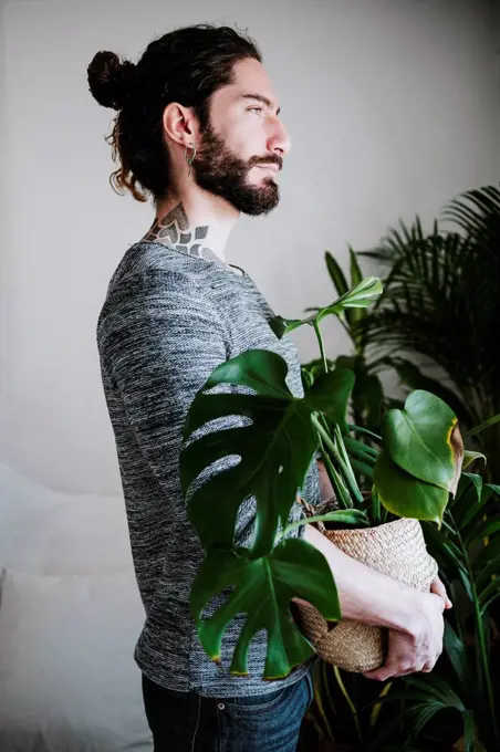 Young man looking away while holding Monstera plant at home